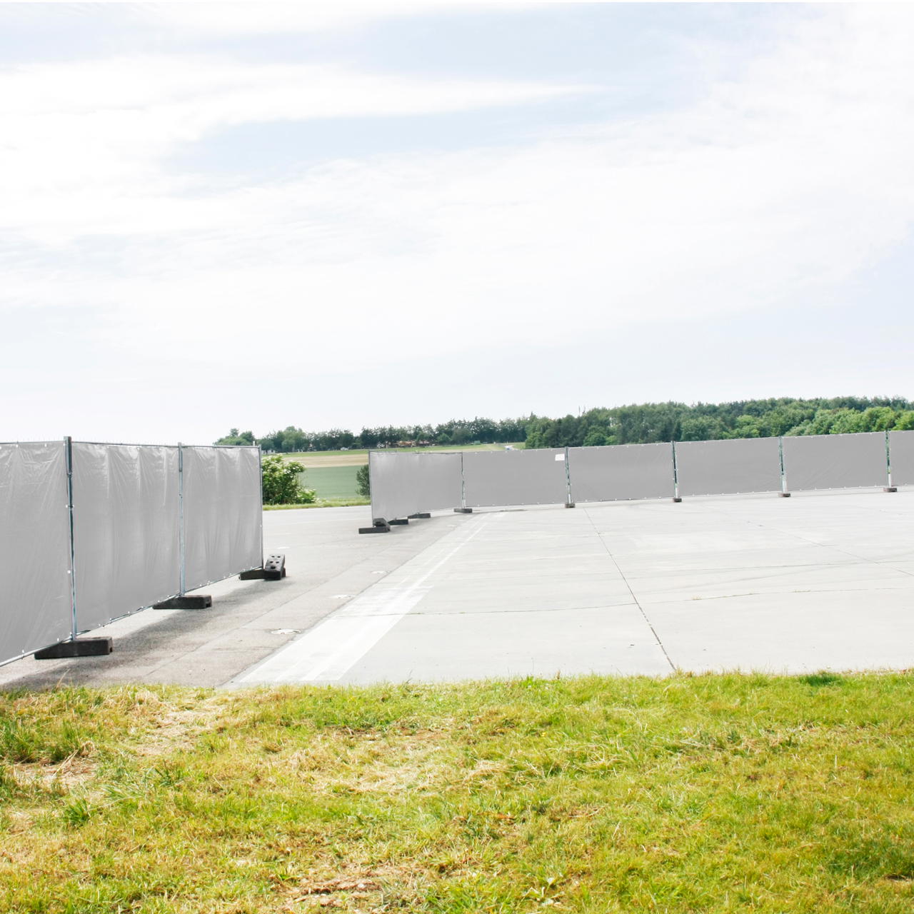 Weiße Gewebeplane als Wind- und Sichtschutz an Bauzaunelementen auf einem Freigelände mit Rasen und befestiger Fläche