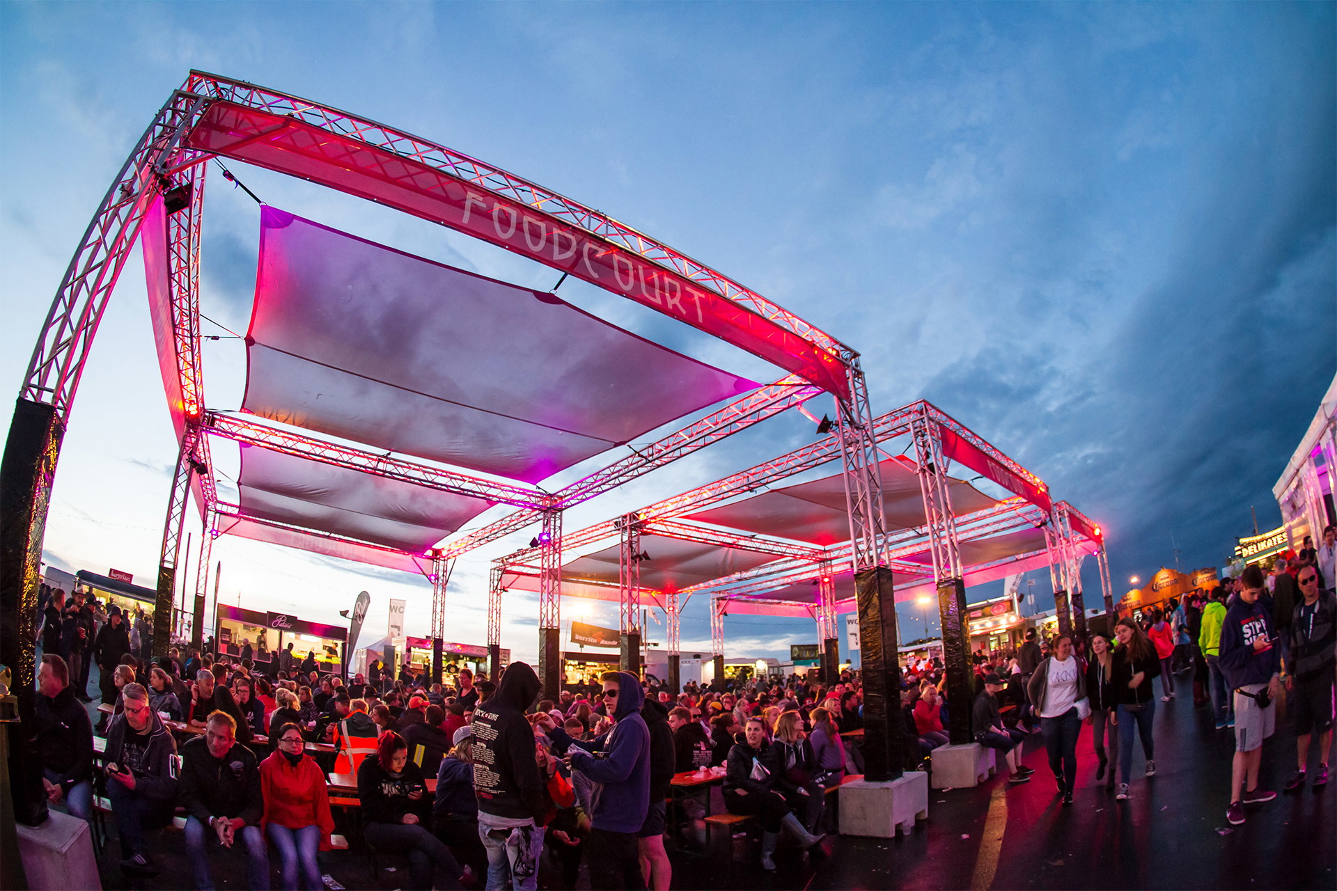Bedrucktes Gastrobanner aus Netzvinyl an Traversenüberdachung über Sitzbereich auf einem Festival bei Abendstimmung