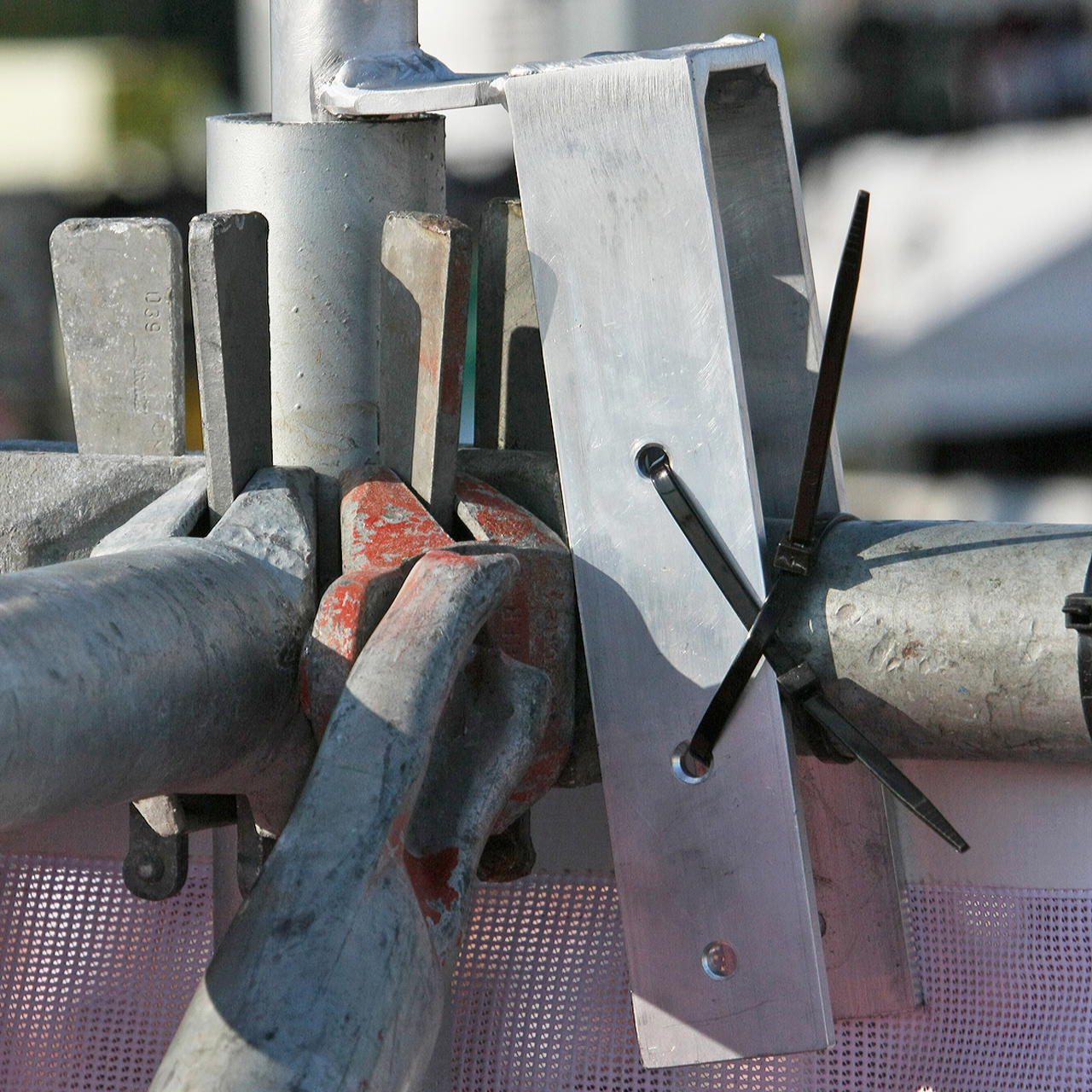 Detailaufnahme des Metalladapters für Bauzaunfahne, über das obere Zaungestänge gesteckt, mit zwei Kabelbindern fixiert