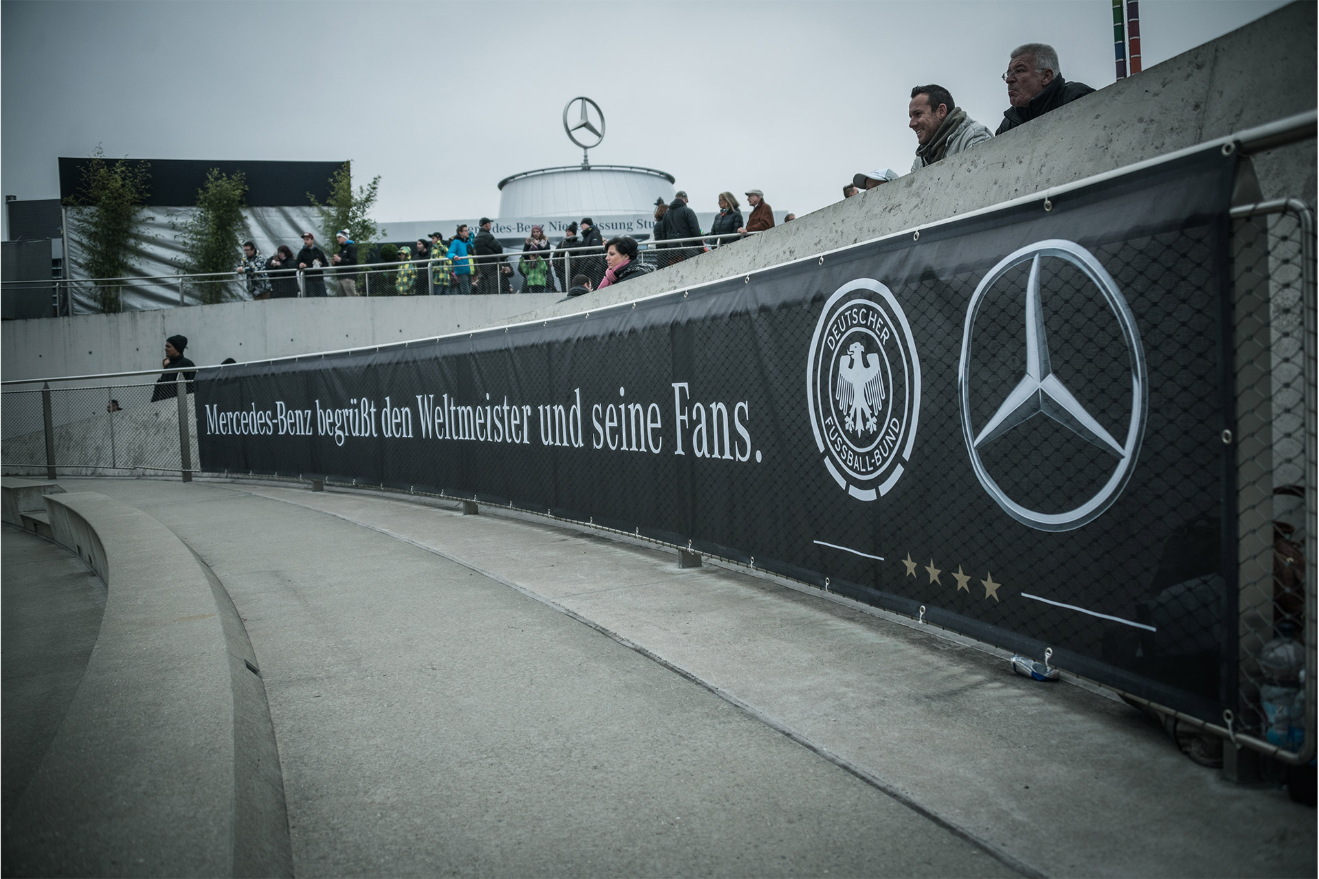 Geöstes Netzvinylbanner bedruckt mit Sponsorenbranding von Mercedes-Benz und DFB entlang eines Zauns mit Zuschauern 