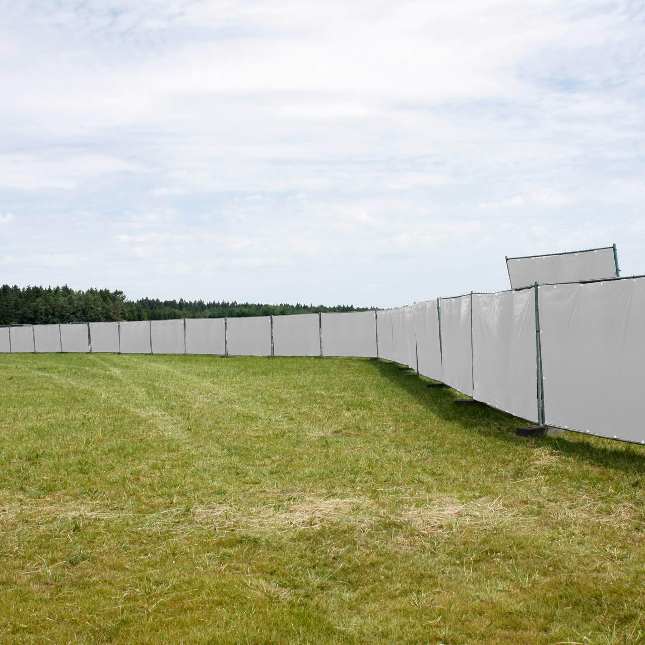 Weiße Bauzaunblenden als Sichtschutz an einer langen, nach links gebogenen Bauzaunreihe auf offenem Rasengelände