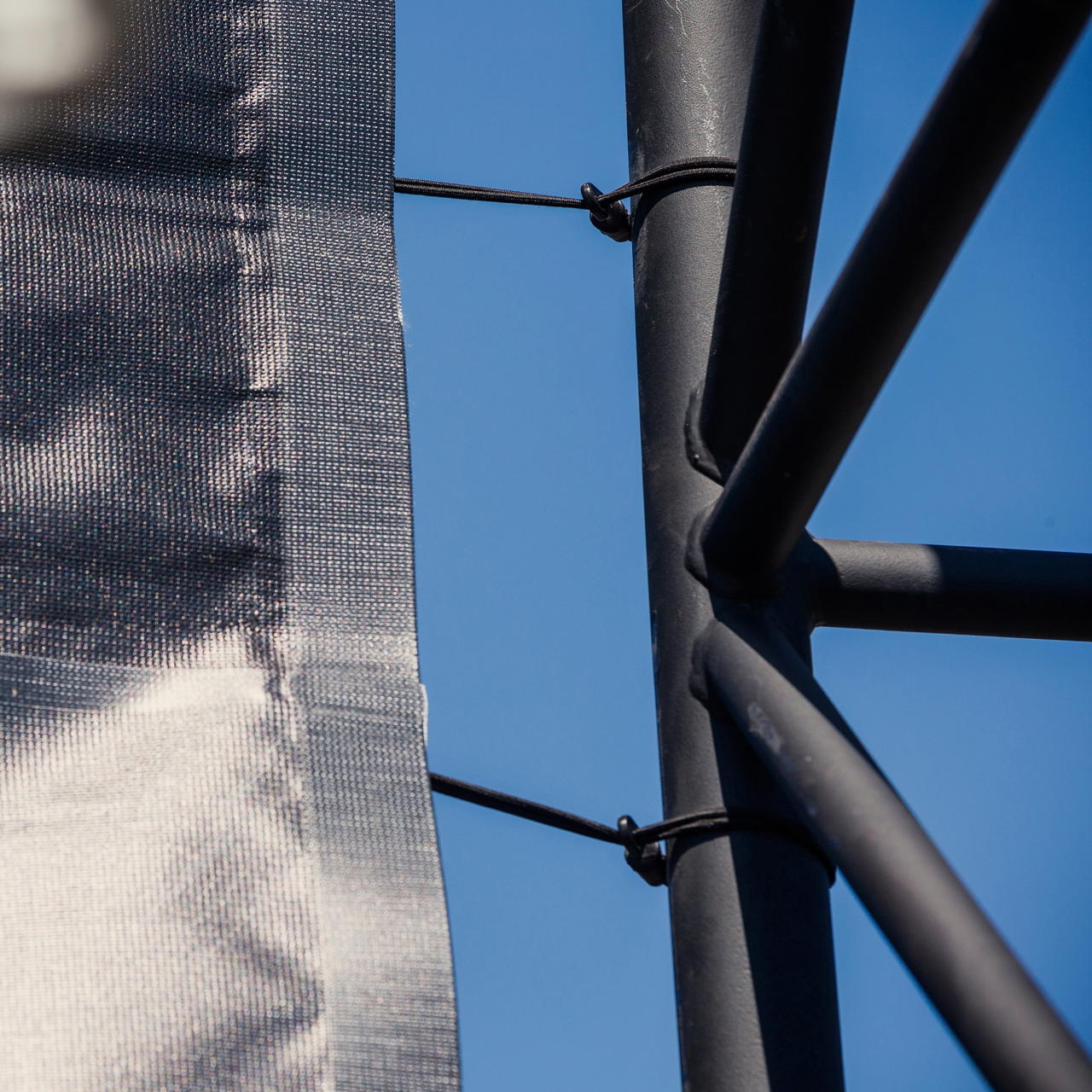 Schwarze Gummispanner in Nahaufnahme zur Befestigung einer Plane an schwarzem Metallgestell vor blauem Himmel