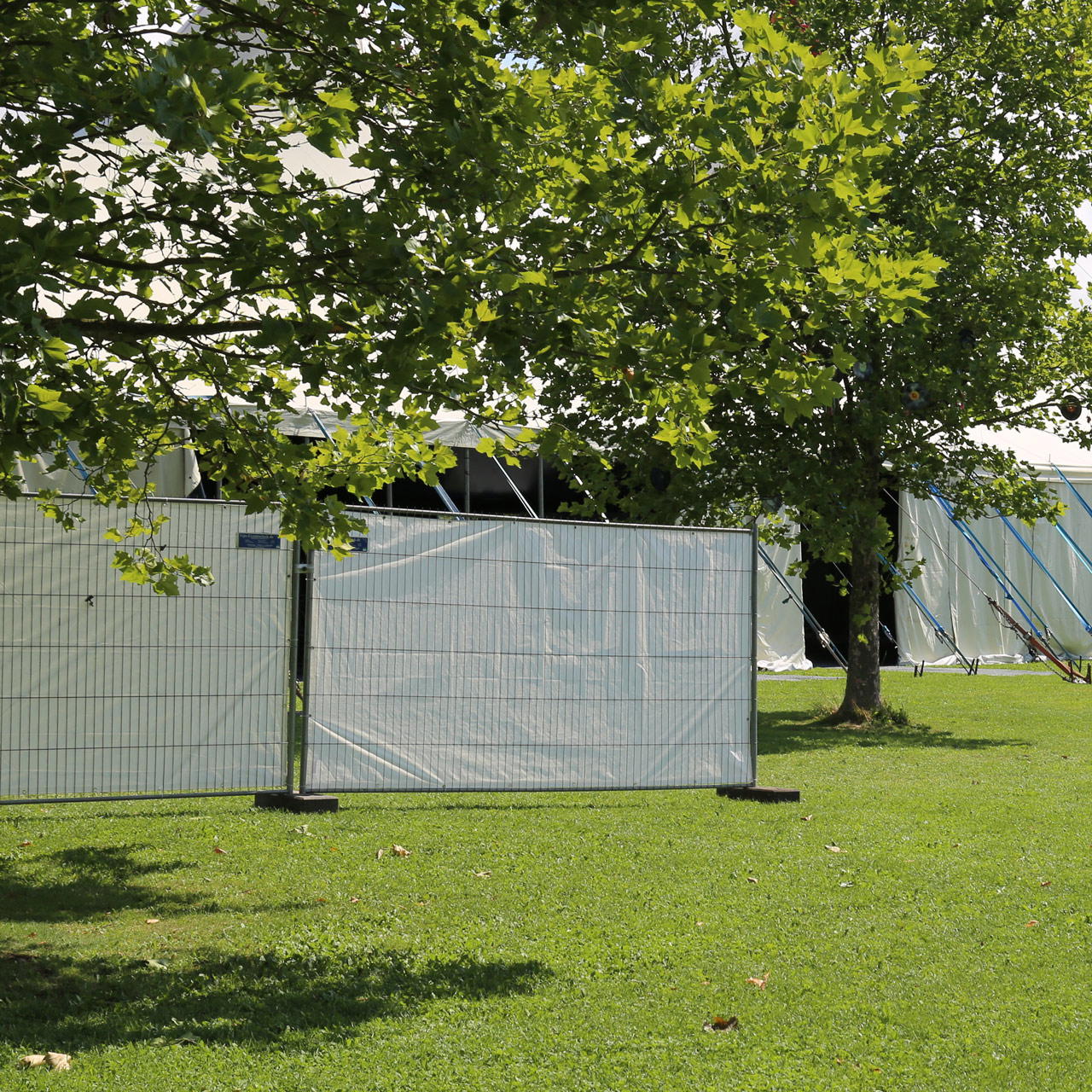 Bauzaunelemente mit weißer Gewebeplane als Sichtschutz auf einer Wiese, aufgestellt vor Eventzelt unter Bäumen