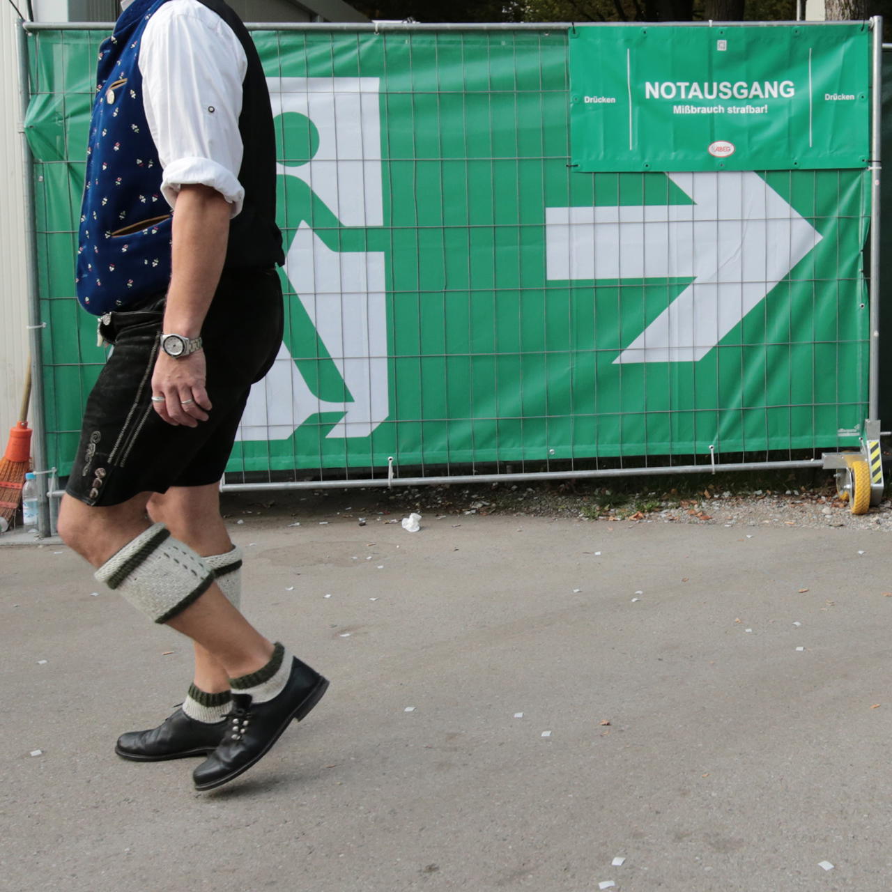 Notausgangsbanner mit Pfeil nach rechts am Bauzaun auf einem Festgelände, ein Mann in Tracht geht daran vorbei