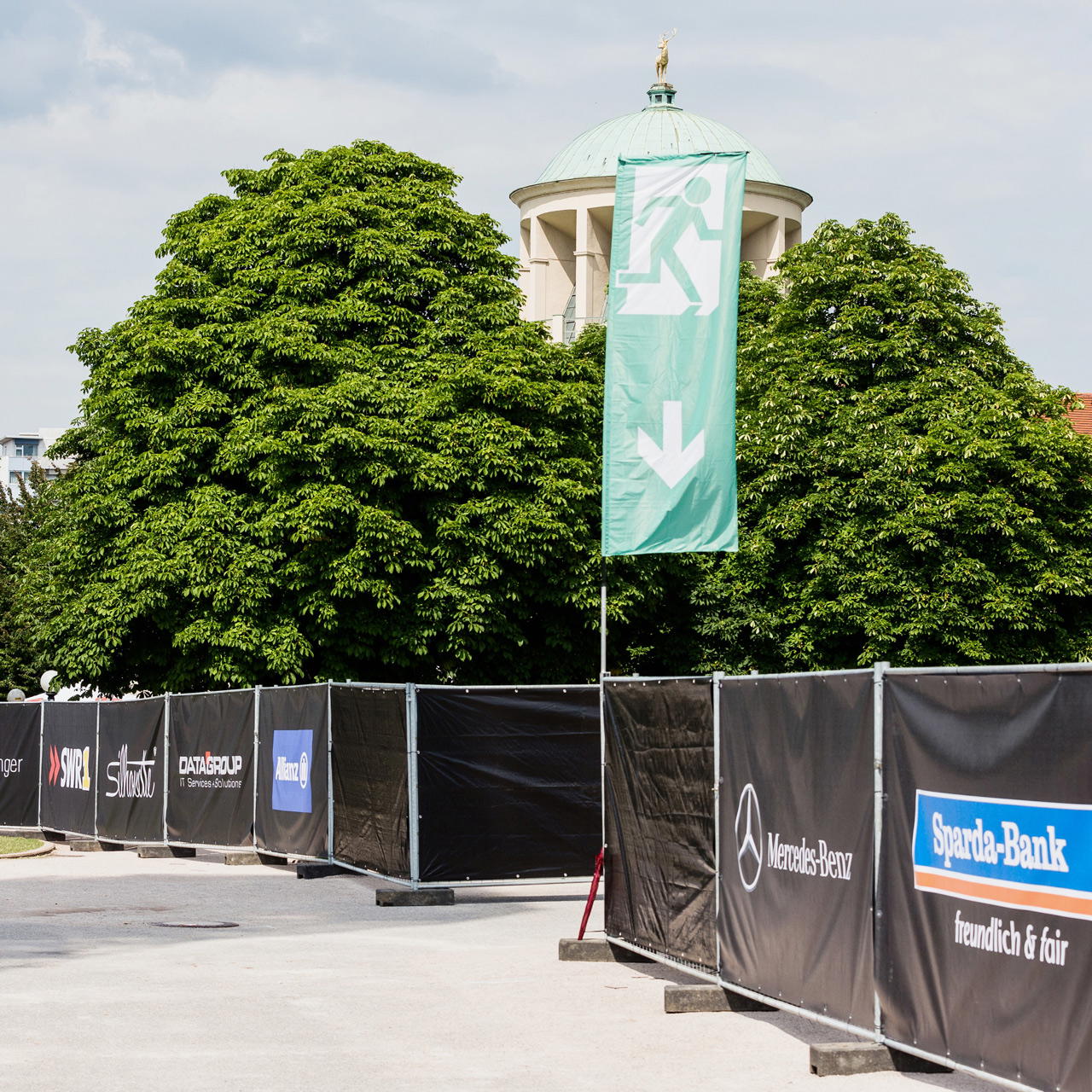 Grüne Flagge mit weißem Notausgangs-Piktogramm und Pfeil nach unten an  einem Mobilzaun mit bedruckten Bauzaunplanen 