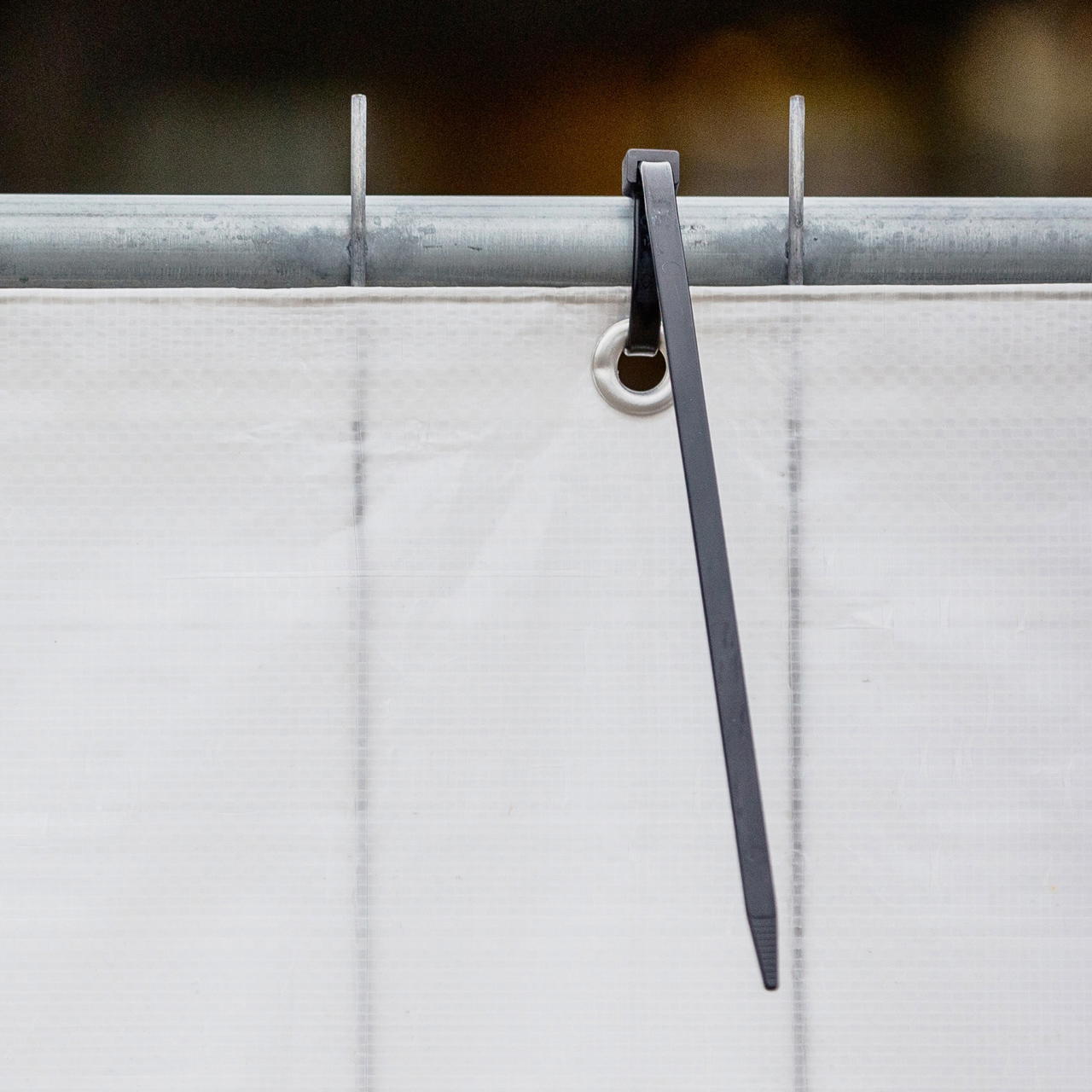 Ein schwarzer Kabelbinder fixiert eine weiße Plane mit Metallöse am oberen Gestänge eines Bauzauns 