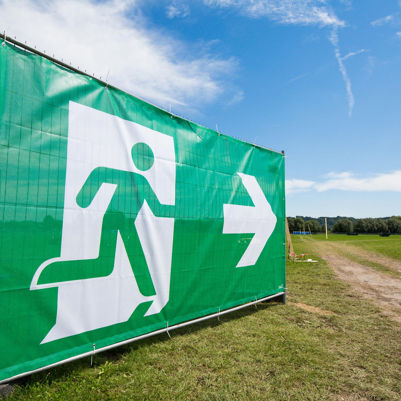 Grünes Notausgangsbanner mit Pfeil nach rechts, befestigt an einem Bauzaun auf einer Wiese bei blauem Himmel