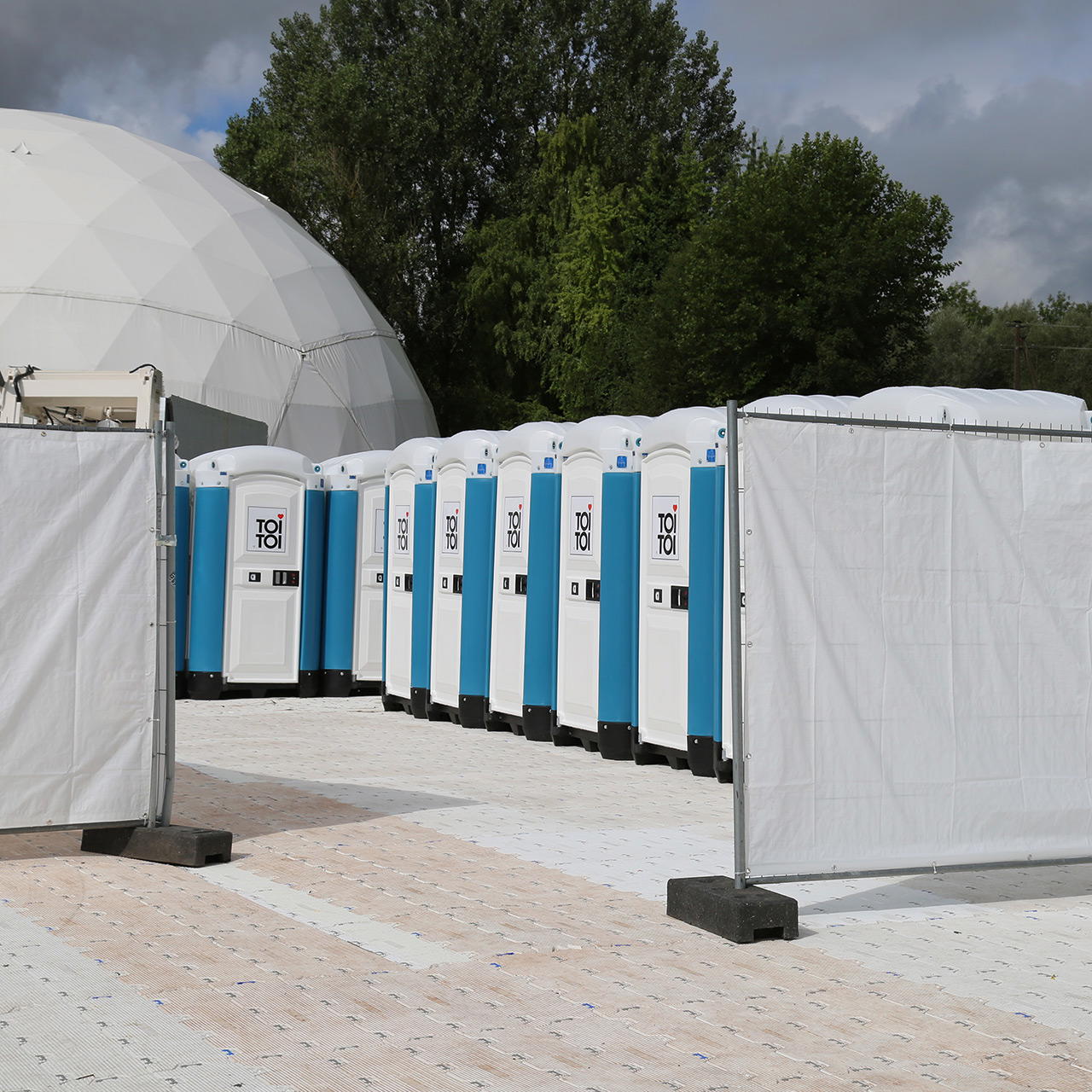 Zwei Bauzäune mit weißen Gewebeplanen bespannt, rahmen den Durchgang zu mobilen Toiletten auf einem Eventgelände