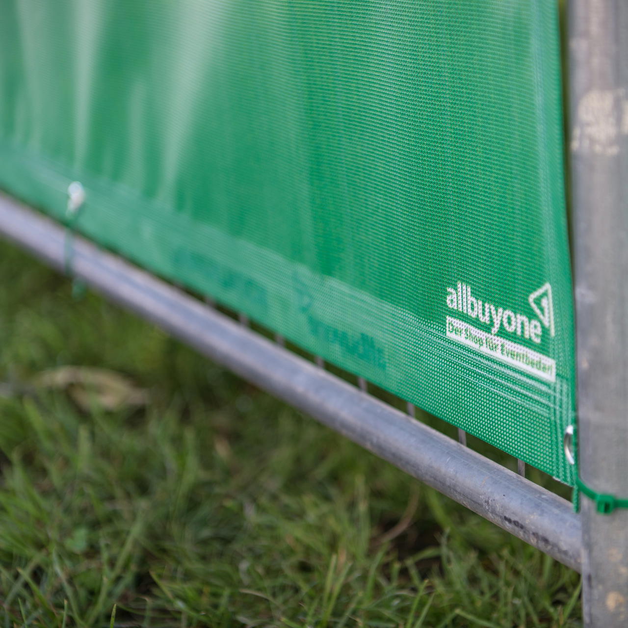 Unterer Bereich eines grünen Meshbanners mit Saum, Öse und weißem Aufdruck, befestigt an einem Bauzaunrahmen