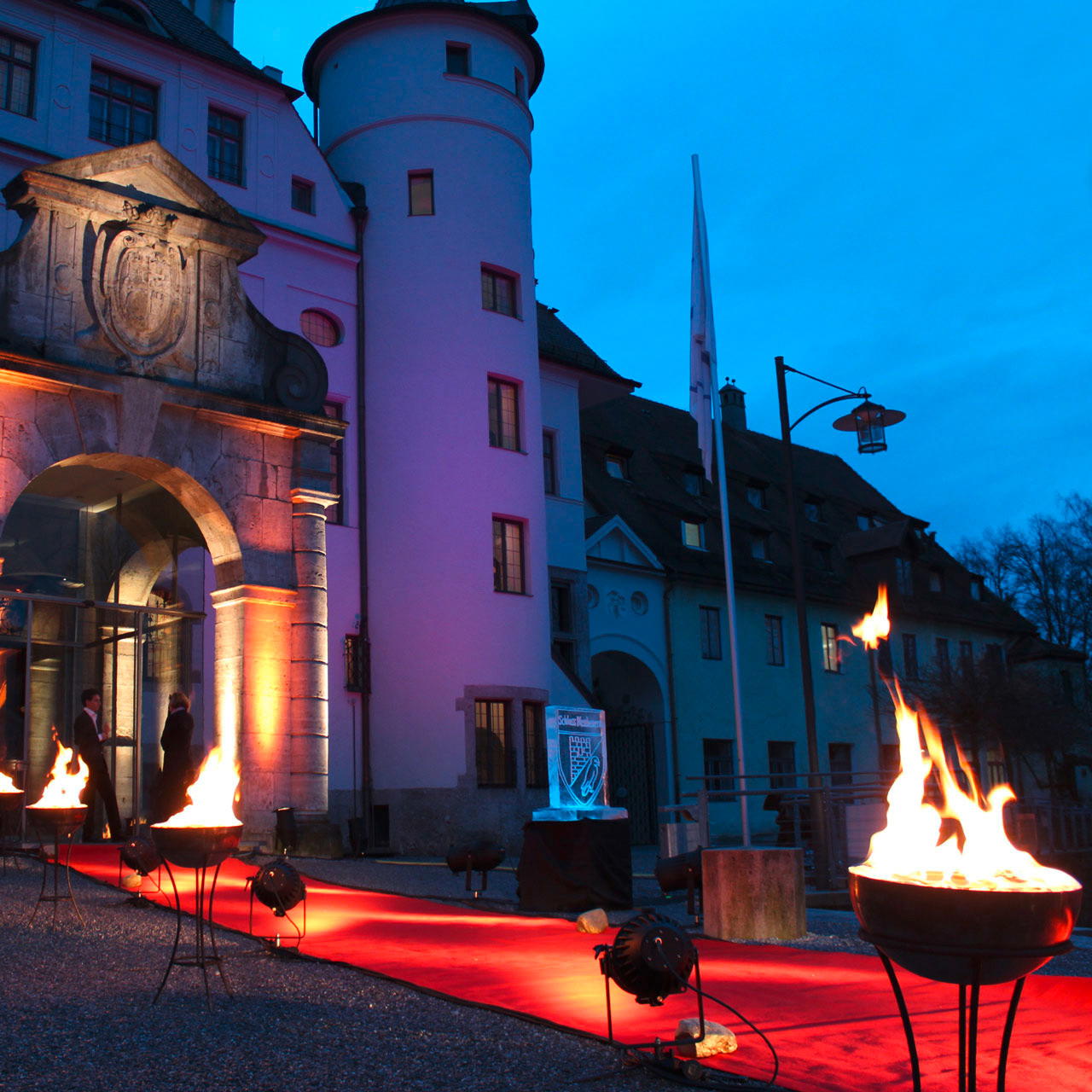 Ausgerollter roter Messeteppich, der auf das Eingangsportal eines Schlosses zuläuft, gesäumt von Feuerschalen