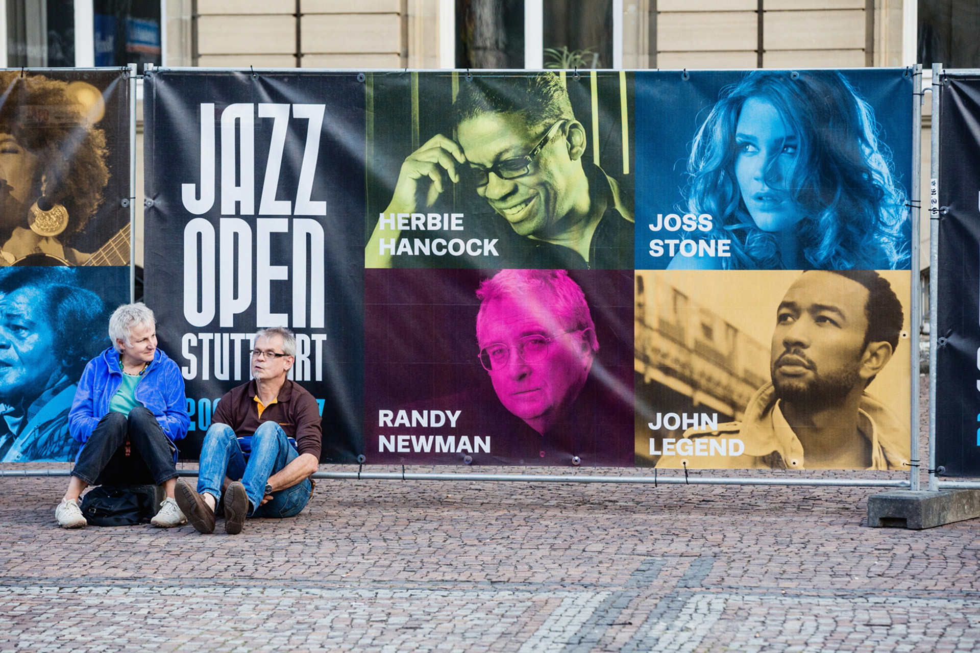 Bauzaun mit bedrucktem Mesh-Banner mit Künstlerporträts und Jazz Open Stuttgart Logo, davor zwei sitzende Personen 