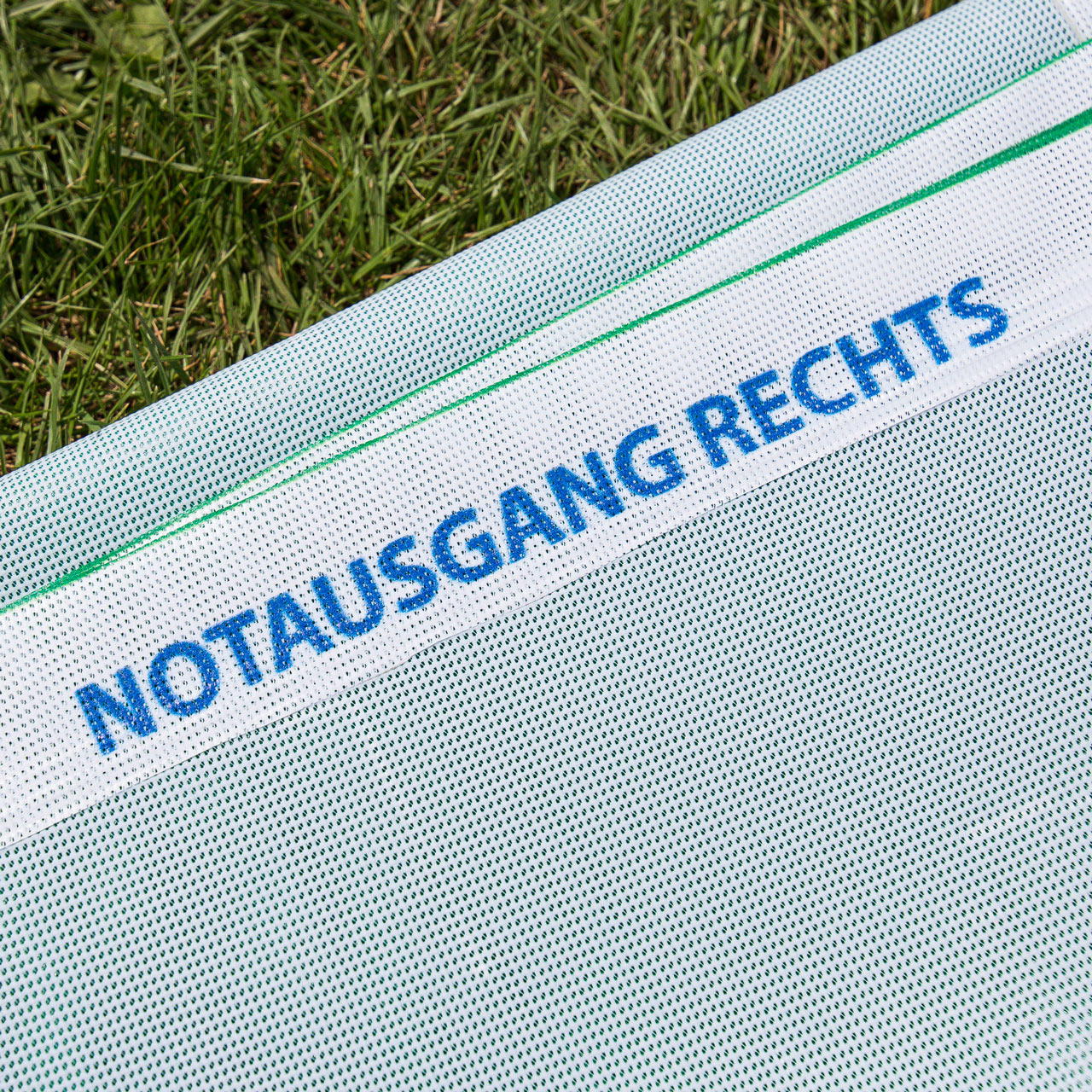 Nahaufnahme der Rückseite eines im Gras liegenden Meshbanners mit Aufdruck „Notausgang rechts“ im weißen Saum