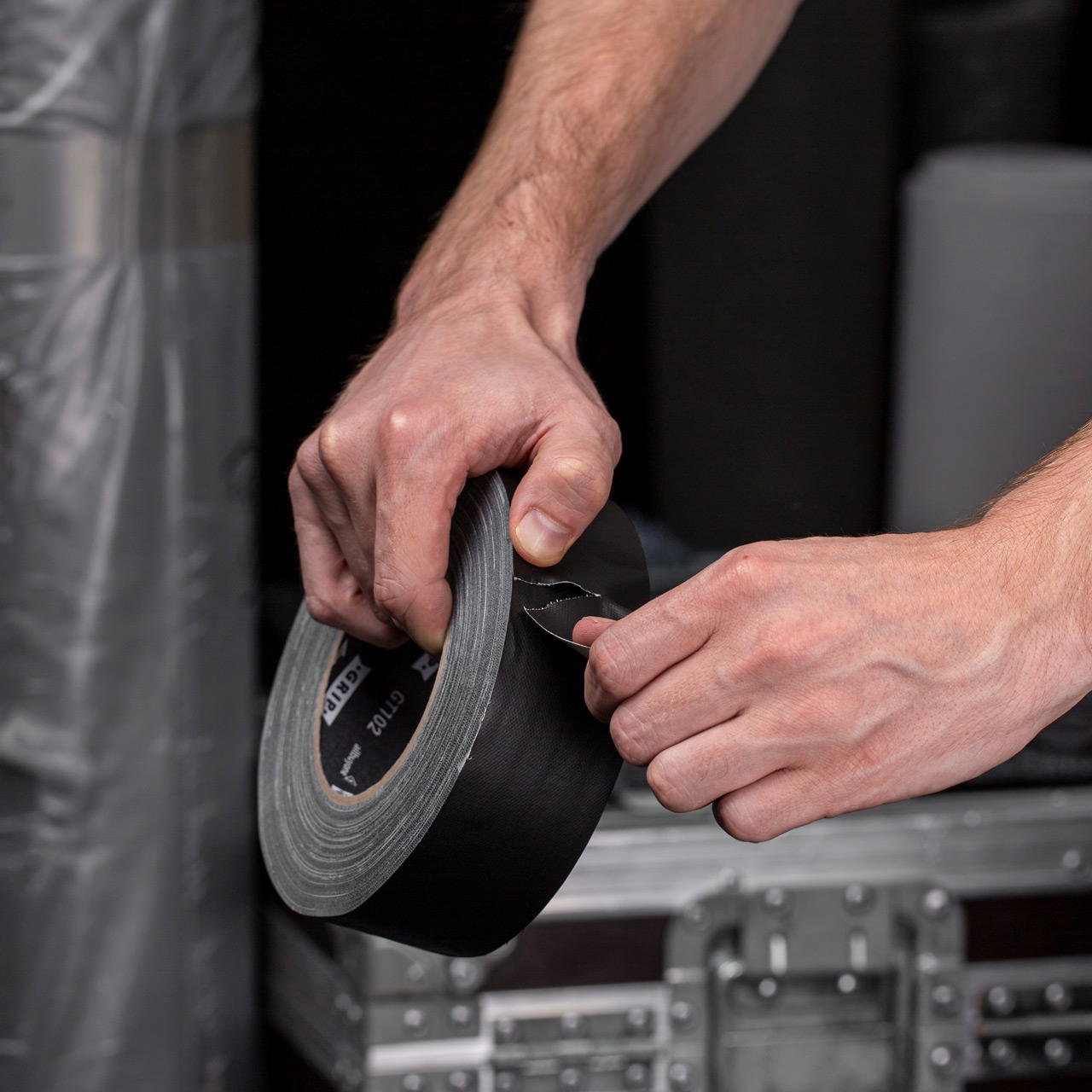 Hände reißen mit sauberer Abrisskante ein schwarzes Gewebeklebeband extra matt ab, im Hintergrund ein Flightcase
