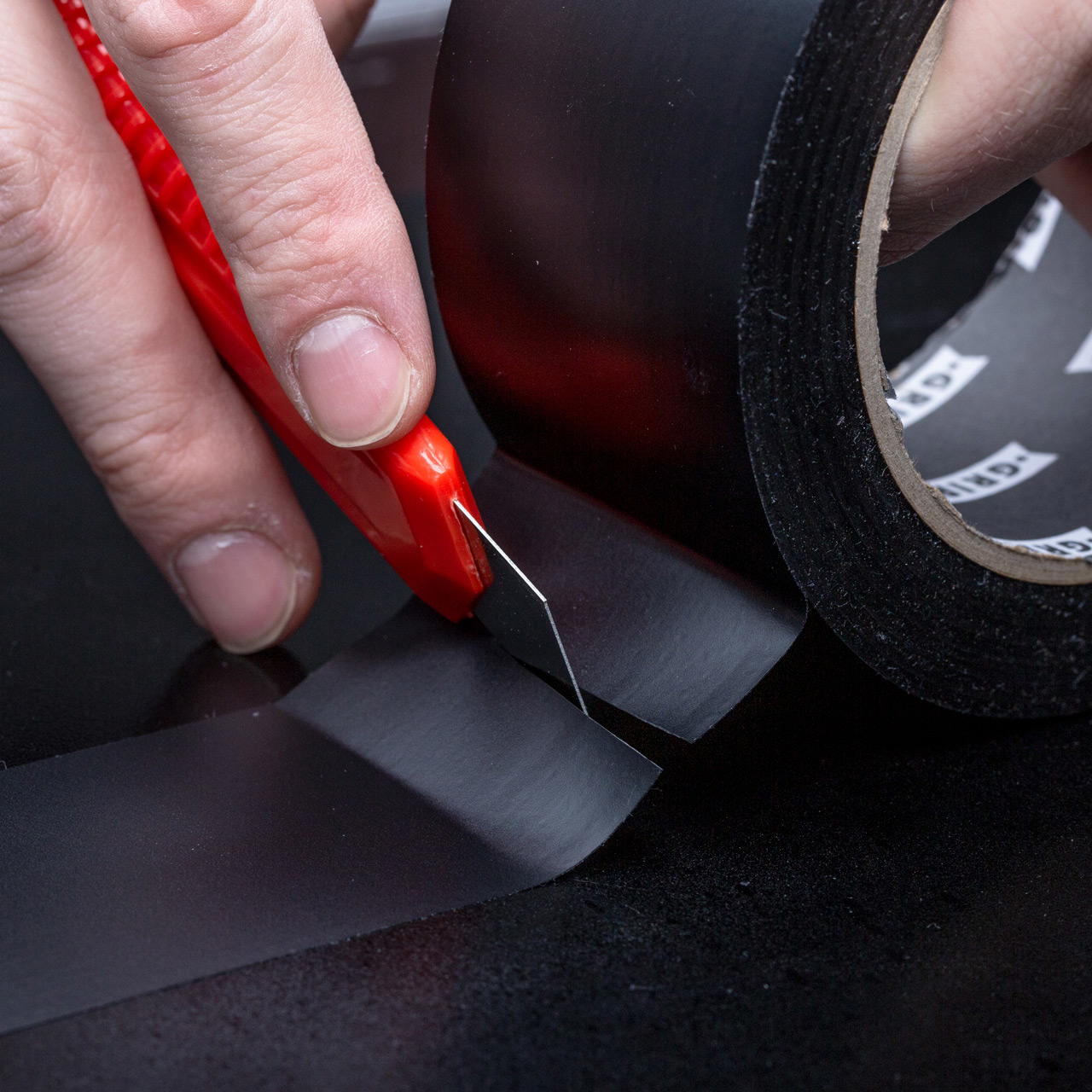 Schwarze Rolle PVC Band mit teilweise auf dunklem Boden verklebtem Band, das mit einem rotem Cutter zugeschnitten wird