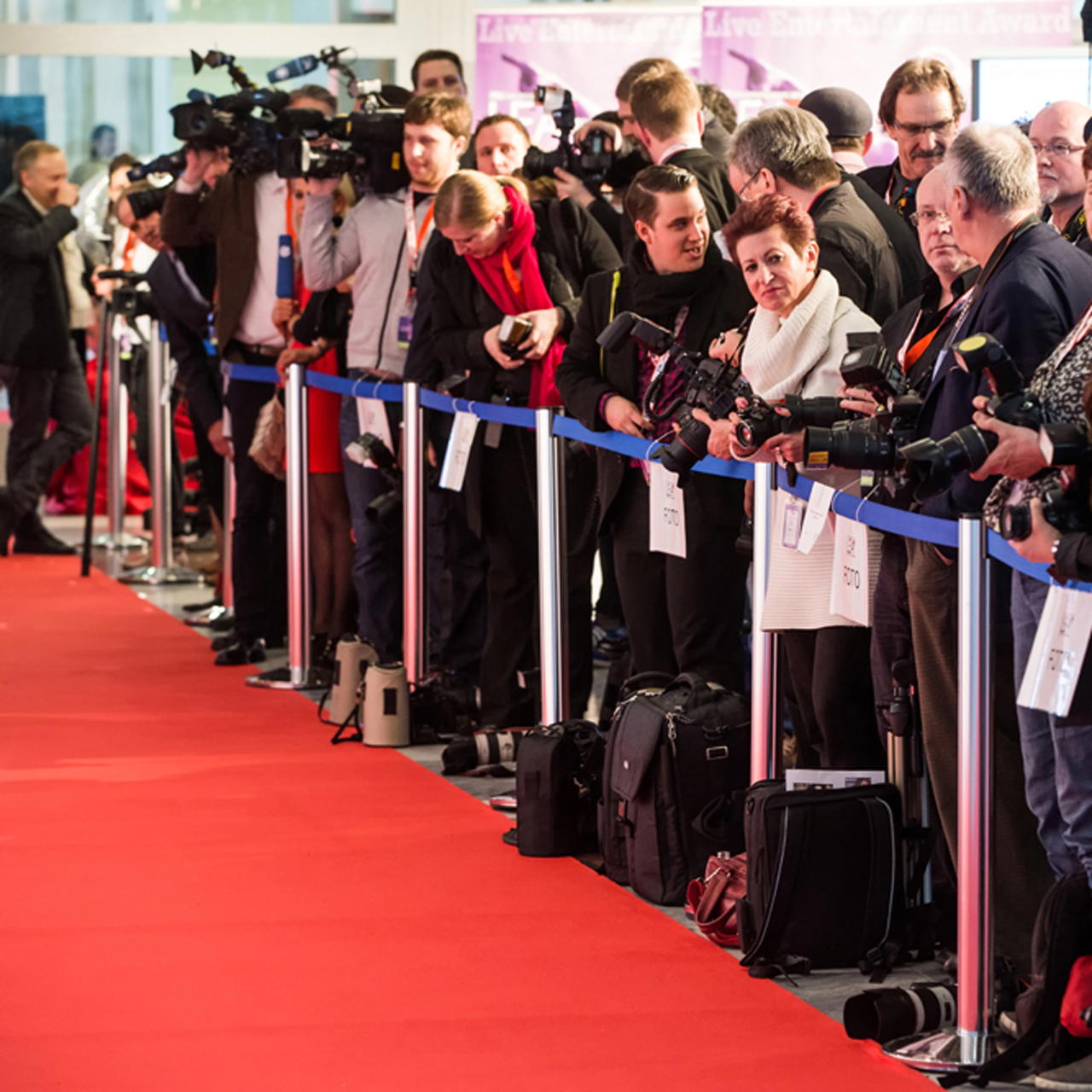 Roter Messeteppich auf einem VIP- Event, gesäumt von Absperrständern, dahinter Fotografen und Presse