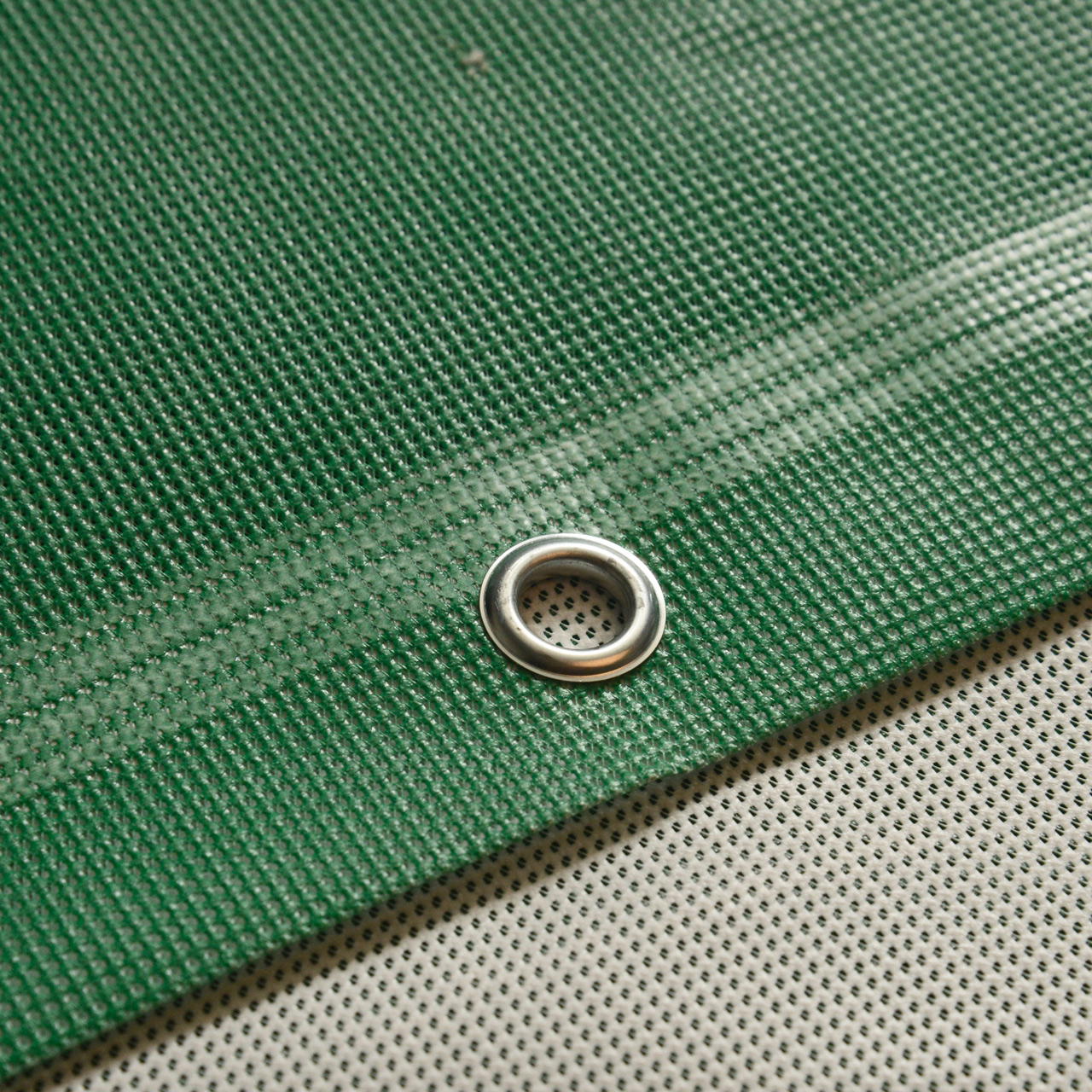 Nahaufnahme eines grünen Meshbanners mit Randverstärkung und Metallöse, liegend auf grauer Rückseite