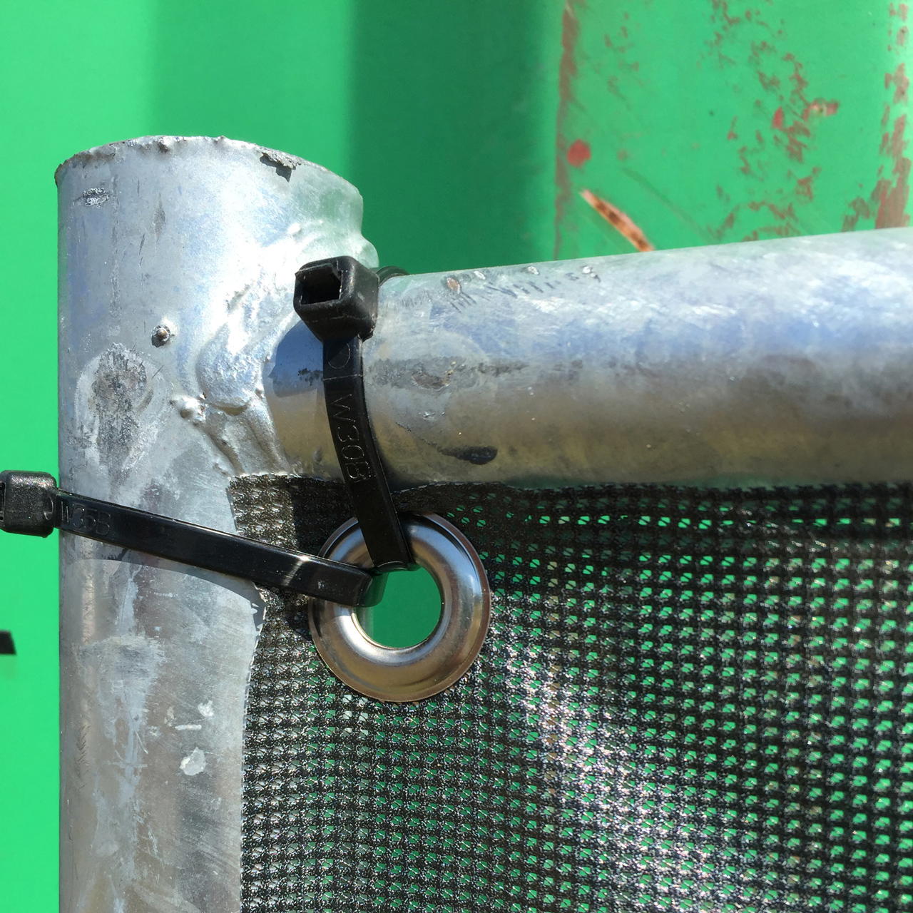 Nahaufnahme von zwei scharzen Kabelbinder, die eine geöste Ecke eines dubkelgrünen Mesh Banners am Bauzaun fixieren 