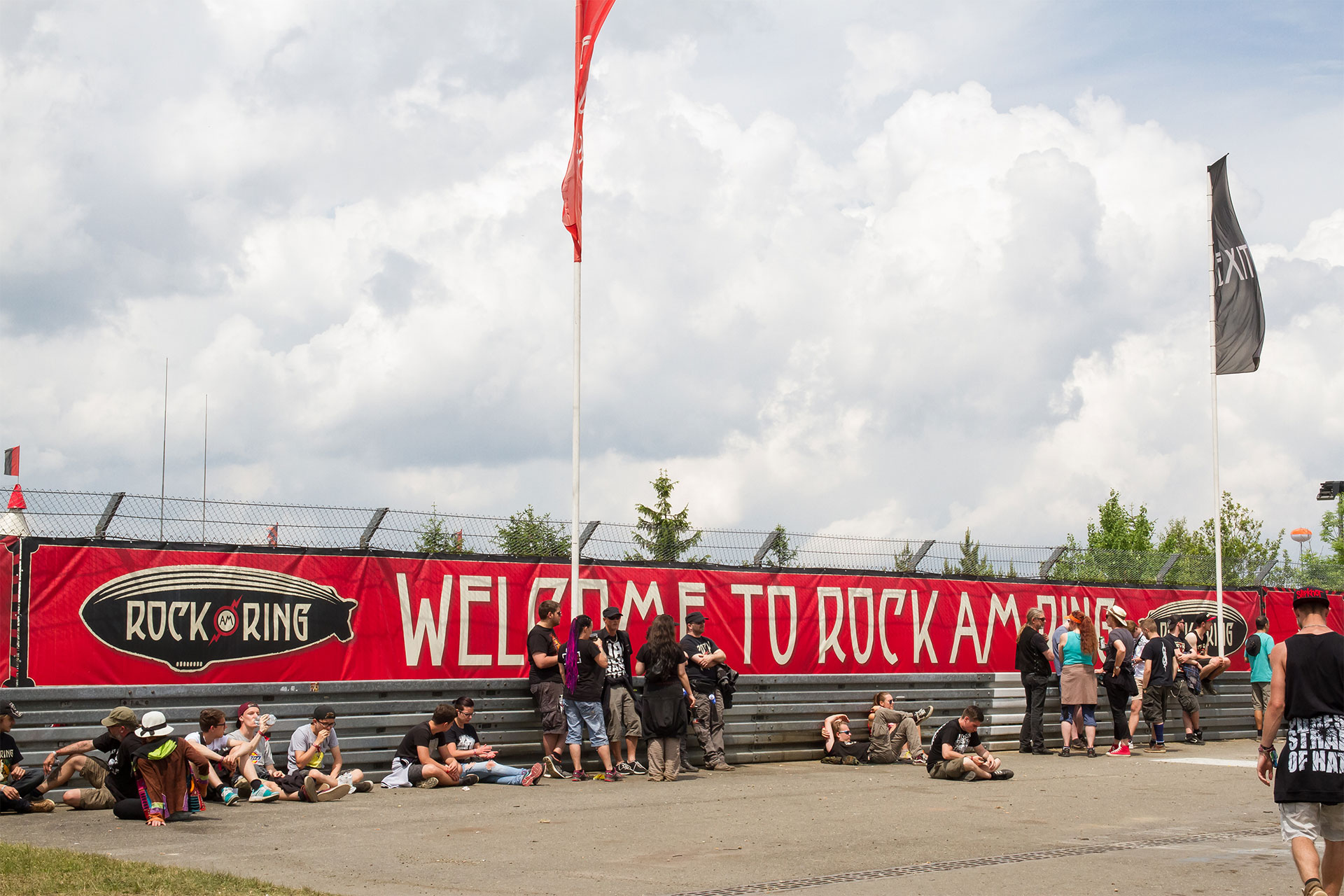 Geöstes rotes Frontlit PVC Banner mit weißem Schriftzug und Logo an einem Zaun mit Festivalbesuchern davor